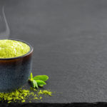 Bowl of blue with matcha green tea, next to it are tea leaves and tea powder on the table. Tea time in the midday heat, the steam rises above the cup, black stone background with copy space , close-up