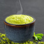 Bowl of blue with matcha green tea, next to it are tea leaves and tea powder on the table. Tea time in the midday heat, the steam rises above the cup, black stone background with copy space , close-up